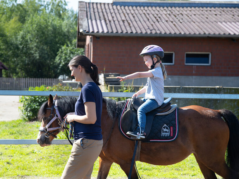 Familienhotel Landhus Laurenz im Münsterland: Mädchen sitzt auf braunem Pferd und wird von Betreuerin geführt
