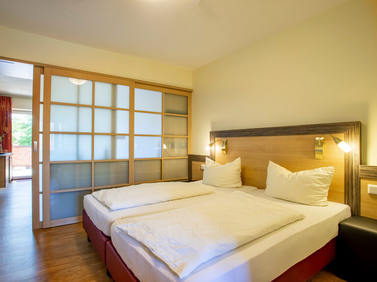 Familienhotel Landhus Laurenz im Münsterland: Doppelbett in Schlafzimmer, das durch Milchglasfront von Wohnbereich abgetrennt ist