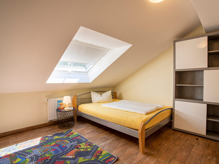 Familienhotel Landhus Laurenz im Münsterland: Kinderzimmer mit Bett und großem Kleiderschrank, Spielteppich am Boden und einer Dachschräge mit Fenster