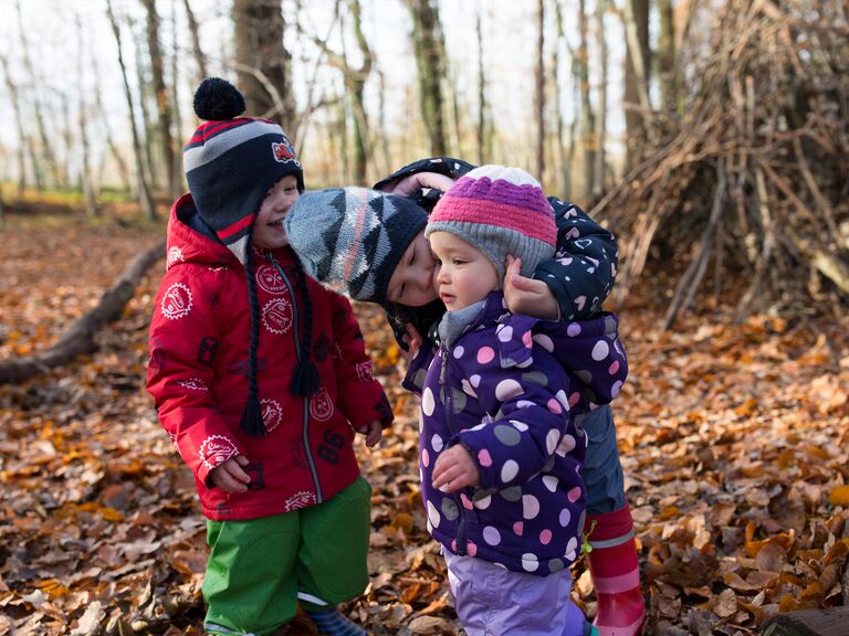 Familienhotel Landhus Laurenz im Münsterland: drei Kinder mit Mützen und in Regenmontur stehen im Herbstwald im Laub. Ein Kind ist zum anderen Kind gebeugt und sagt etwas.